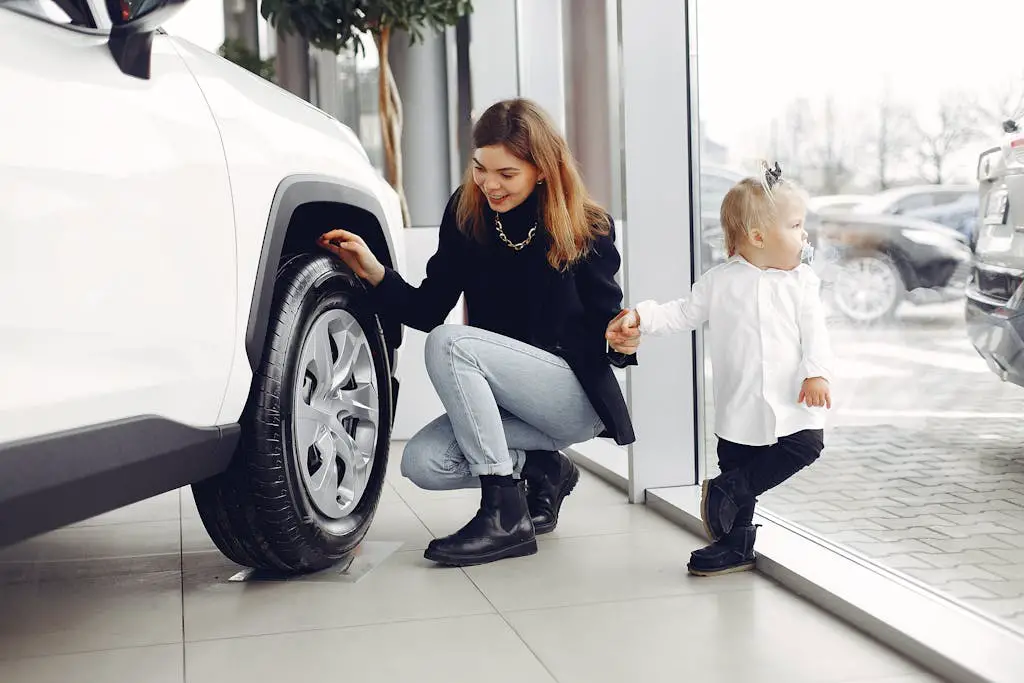 Full length of cheerful smiling mother in jeans holding hands with cute little girl and looking at car wheel in car showroom in daylight